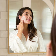 Woman applying cream to her face in front of a mirror