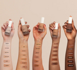 Five hands holding bottles of foundation with swatches on a beige background