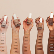 Five hands holding bottles of foundation with swatches on a beige background
