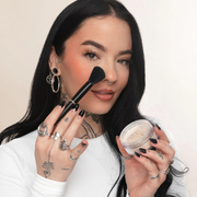 Woman applying makeup with a brush and compact powder against a neutral background