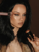 Woman applying makeup with a brush and compact mirror, wearing a fur-lined garment.